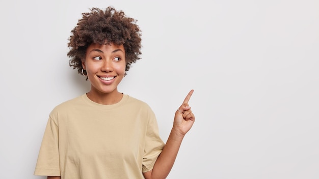 Foto il colpo orizzontale di una donna di bell'aspetto con l'indice dei punti di capelli ricci più fine sopra al banner promozionale suggerisce che la visita al negozio mostra uno spazio vuoto per la copia su sfondo bianco. concetto pubblicitario