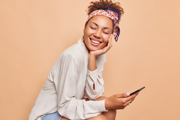 Horizontal shot of pretty millennial ethnic woman smiles happily keeps eyes closed browses mobile phone wears shirt and kerchief tied over head isolated over beige wall