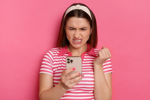 Horizontal shot of sad irritated young girl with hair band wearing striped casual t shirt holding with smart phone in hands reading crazy message expressing anger standing isolated pink background