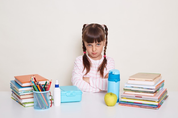 Horizontal shot of sad little schoolgirl with dark hair and braids sitting at table surrounded with books looking at camera with pout lips and upset eyes expressing sadness and sorrow