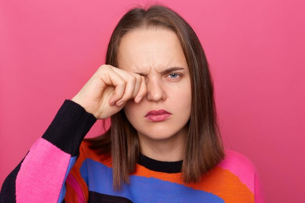 Horizontal shot of sad tired stressed woman wearing colorful jumper posing isolated over pink background looking at camera and crying rubbing her eye feels pain