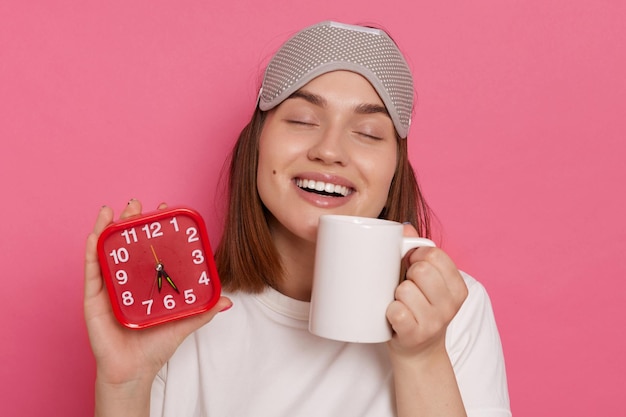 Horizontal shot of satisfied adorable woman with sleeping mask holding cup with coffee and coffee cup waking up with good mood enjoying her morning beverage