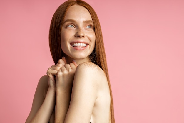 Horizontal shot of satisfied caucasian woman clenching fists with pleasure, looking aside with happy expression, broadly smiling isolated over pink background. Body care, spa, emotions concept.