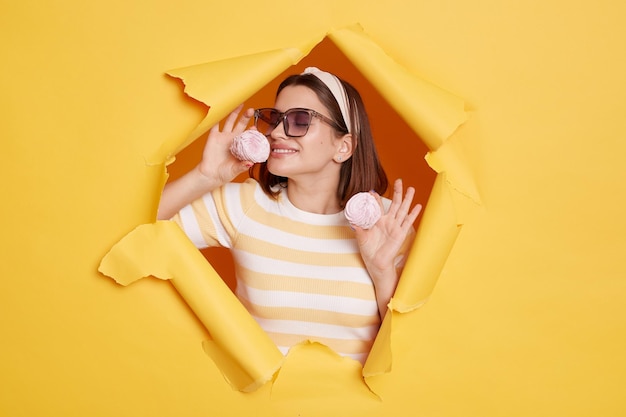 Horizontal shot of satisfied delighted woman wearing hair band and striped t shirt standing in yellow paper hole smelling marshmallow and wants to eat sugary dessert