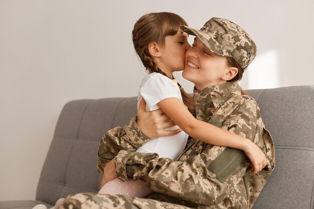 Horizontal shot of satisfied little girl with m other military\
woman wearing camouflage uniform and cap posing with her daughter\
family hugging each other and expressing happiness