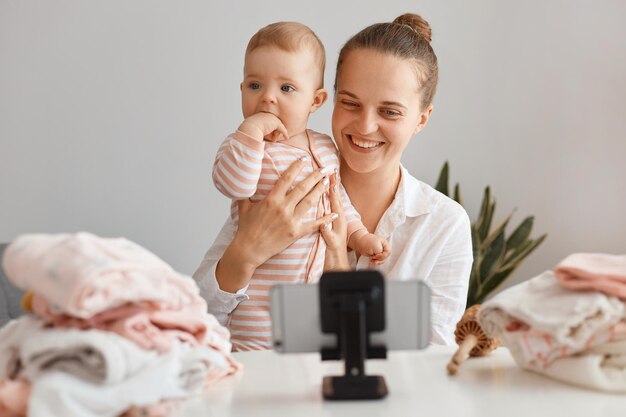 Horizontal shot of satisfied woman blogger sitting at table with toddler kid and recording video for her vlog, making content with her daughter, having pleasant conversation with subscribers.