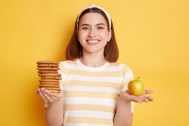 新鮮なリンゴと黄色の背景で隔離のおいしい甘いクッキーを保持しているカジュアルなスタイルの服を着て黒髪で満足している若い女性の水平方向のショット