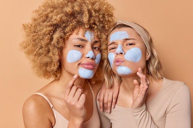 Horizontal shot of serious attentive women examine condition of\
facial skin apply clay mask take care of complexion stands closely\
to each other isolated over brow background beauty treatments