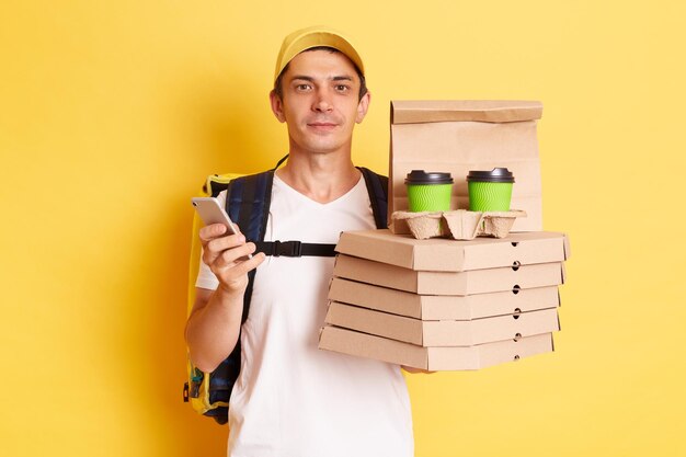 Horizontal shot of serious calm man courier with thermo backpack in white Tshirt and cap looking at camera holding client's order standing with smart phone in hands isolated over yellow background