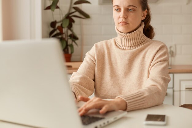 Horizontal shot of serious concentrated Caucasian woman wearing beige jumper posing in kitchen and working on notebook typing on keyboard working online