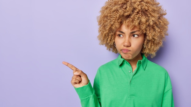 Horizontal shot of serious displeased woman with curly hair\
purses lips points away on blank space shows something unpleasant\
dressed in green jumper isolated over purple background look at\
this