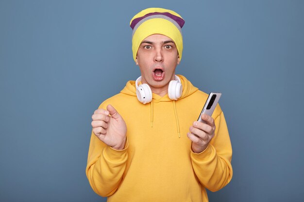 Horizontal shot of shocked amazed man wearing beanie hat and yellow hoodie holding smart phone in hands reading breaking new in internet looking at camera with open mouth posing against blue wall