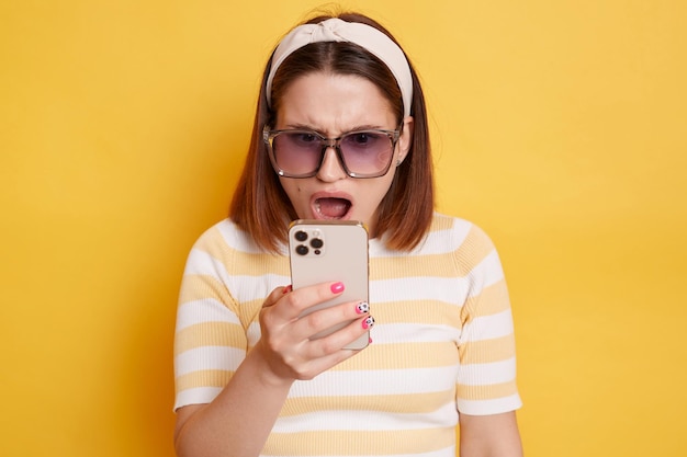 Horizontal shot of shocked astonished woman wearing striped t shirt and sunglasses holding smartphone in hands looking at display with open mouth posing isolated over yellow background
