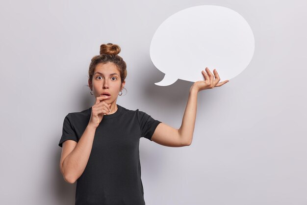 Photo horizontal shot of shocked european woman with hair bun stares impressed hold empty communication bubble dressed in casual black t shirt isolated over white background place your text here