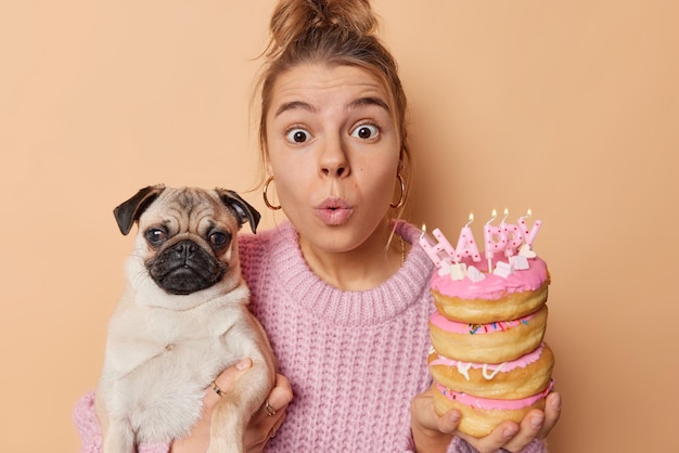 Horizontal shot of shocked young woman stares stupefied at camera keeps lips rounded poses with pug dog and pile of doughnuts wears knitted sweater isolated over brow background Animals concept