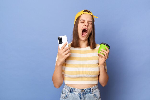 Horizontal shot of sleepy tired teen girl wearing striped Tshirt and baseball cap standing isolated over blue background holding coffee to go and cell phone yawning needs energy