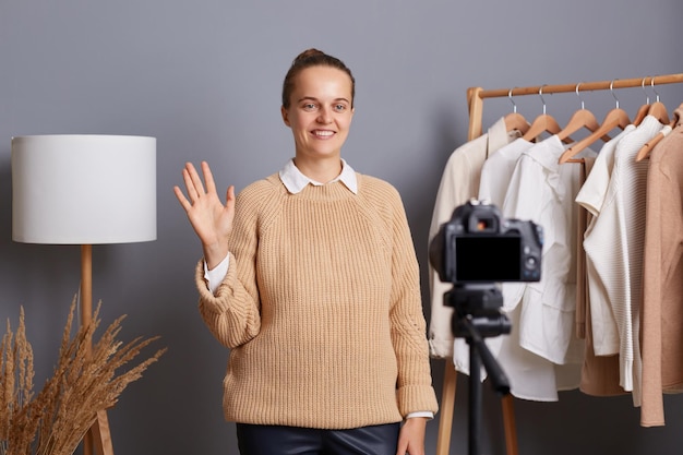 Horizontal shot of smiling beautiful caucasian wearing beige\
jumper recording video in her showroon for her blog about fashion\
waving hand saying hello to followers