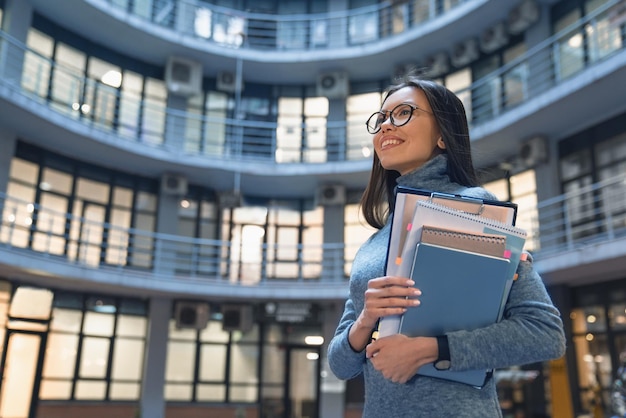 Photo horizontal shot of smiling business woman standing outside office building with her work accessories in arms confident and cheerful successful female manager in the office yard