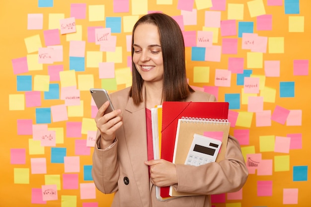 Horizontal shot of smiling cheerful woman wearing beige jacket\
standing against yellow wall covered with stickers holding paper\
folders with documents using cell phone