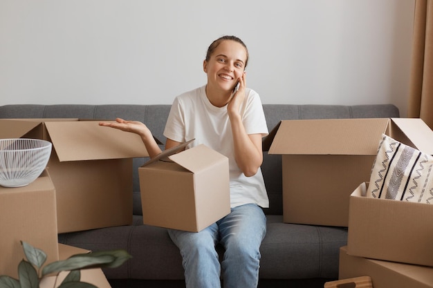 Photo horizontal shot of smiling confused woman wearing white casual t-shirt and jeans, sitting on sofa, surrounded with boxes during moving, talking phone and spreading arm.