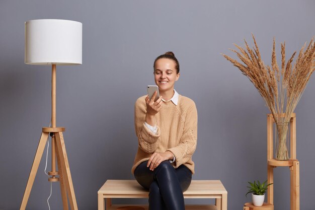 Horizontal shot of smiling delighted woman wearing casual outfit posing against gray wall with lamp and dried flower on background sitting on wooden bench using mobile phone