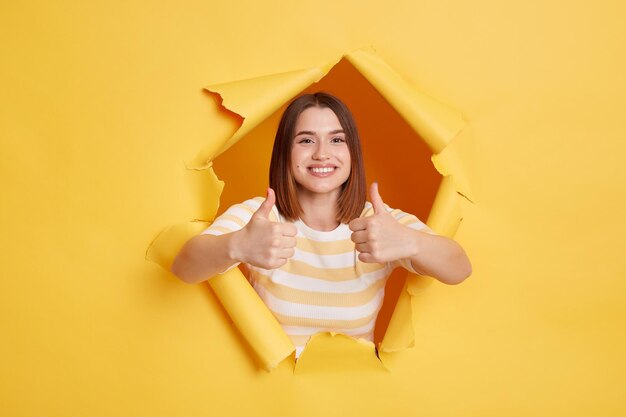 Horizontal shot of smiling happy woman wearing striped t shirt looking through yellow paper torn showing thumb up approval gesture good feedback