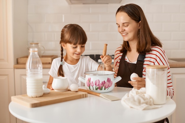 Horizontal shot of smiling mother in apron sitting at table with daughter and cooking together in kitchen mixing dough for homemade pastry expressing happiness