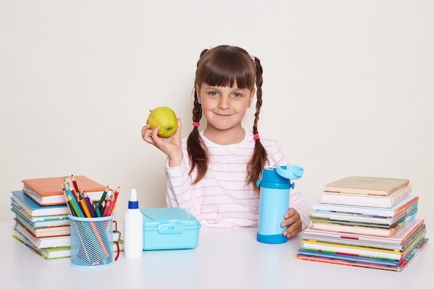 Horizontal shot of smiling optimistic little schoolgirl with pigtails wearing striped shirt sitting at the desk surrounded with books and other school supplies holding fresh fruit looking at camera