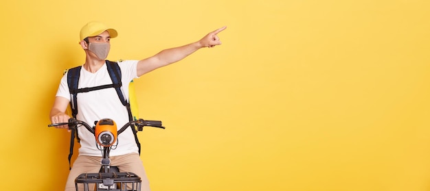 Horizontal shot of smiling positive delivery man in protective mask with thermo backpack pointing at advertisement area delivering orders on electric scooter isolated on yellow background