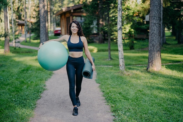 Horizontal shot of sportswoman wears cropped top and leggings carries fitness ball and rolled up karemat walk on road around trees and green grass leads healthy lifestyle Sport equipment