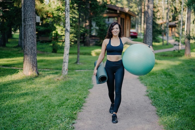 Horizontal shot of sportswoman wears cropped top and leggings carries fitness ball and rolled up karemat walk on road around trees and green grass leads healthy lifestyle Sport equipment