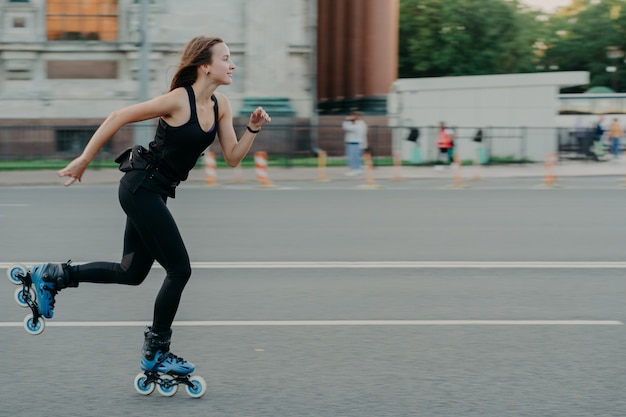 Horizontal shot of sporty energized woman enjoys rolleblading being photographed in motion poses on road against blurred street background has regular fitness activities for keeping fit and healthy