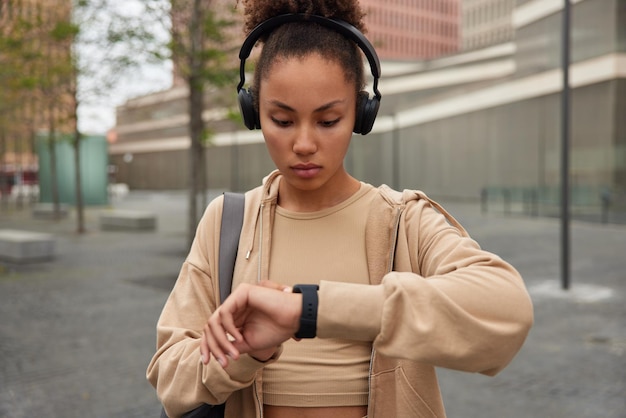 Foto l'inquadratura orizzontale di una donna sportiva controlla il tempo sullo smartwatch moderno focalizzato sul display ascolta la musica preferita tramite cuffie wireless vestite con abbigliamento sportivo posa all'aperto su sfondo sfocato