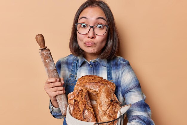 Horizontal shot of surprised asian housewife purses lips holds\
rolling pin and homemade freshly baked bread wears transparent\
eyeglasses and blue shirt isolated over beige background bakery\
food