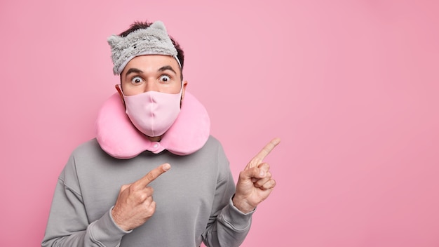 Horizontal shot of surprised European man wears protective face mask against coronavirus blindfold neck pillow indicates at blank space