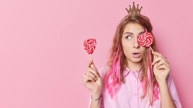 Horizontal shot of surprised young female pretends being princess holds two lollipops keeps mouth opened looks wondered away poses against pink background blank copy space for your promotion