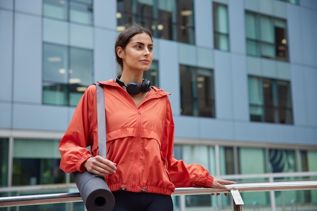 Horizontal shot of thoughtful brunette sporty woman dressed in sportswear carries rolled karemat focused into distance stands against blurred modern building prepares for workout or yoga practice