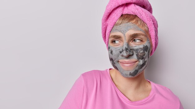 Horizontal shot of thoughtful woman applies beauty clay mask on\
face focused aside undergoes skin care procedures wears pink t\
shirt nd bath towel wrapped on head isolated over grey\
background