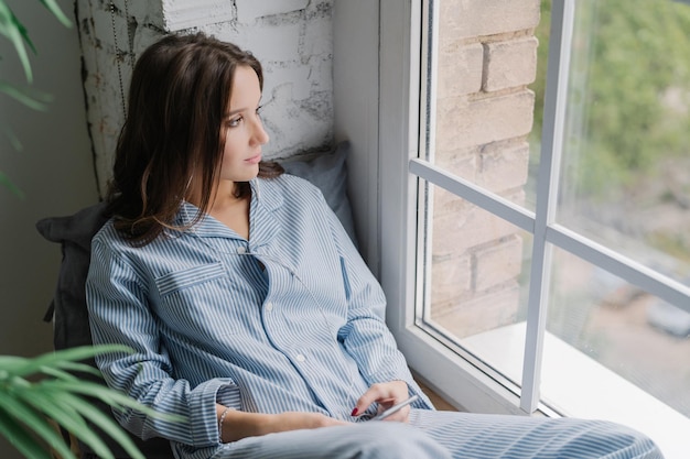 Horizontal shot of thoughtful young woman sits on windowsill
dressed in casual nightwear uses smart phone and earphones listens
calm music while looks thoughtfully in window enjoys home time