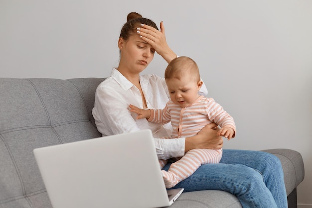 Horizontal shot of tired exhausted female with bun hairstyle wearing white shirt and jeans sitting on sofa with her toddler daughter, being exhausted work online via laptop and looking after baby.