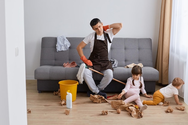 Horizontal shot of tired exhausted man with terrible pain in neck sitting on sofa and touching back having rest while washing floor in room where his children playing