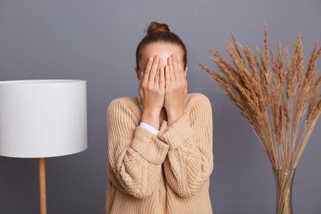Horizontal shot of unknown anonymous female in beige sweater sitting near lamp at home in living room covering her eyes with palms hiding her face behind her hands