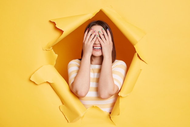 Horizontal shot of unknown anonymous woman stands in torn paper hole posing and covering eyes with palms looking through breakthrough of yellow background