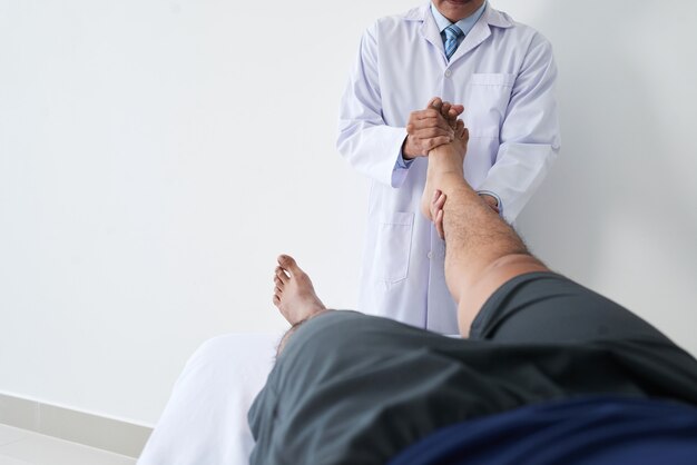 Горизонтальный снимок неузнаваемого врача-остеопата, работающего с пациентом мужского пола, держащим ногу и выполняющим манипуляции с суставом стопы