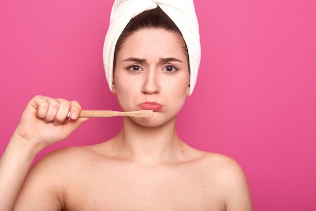 Horizontal shot of upset woman holding toothbrush and brushing her teeth isolated over pink