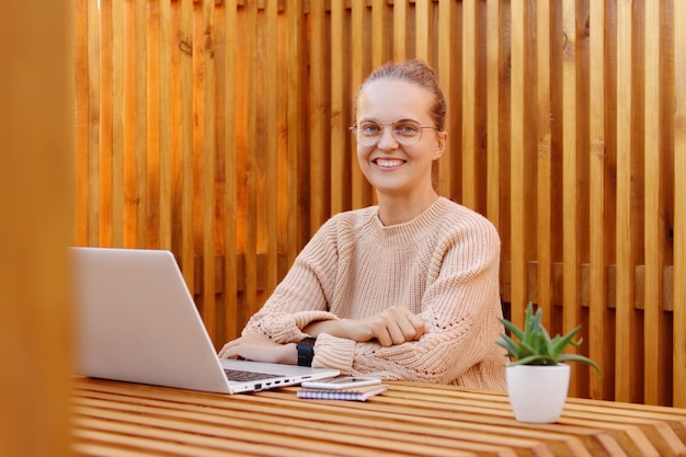 Horizontal shot of winsome adorable woman with bun hairstyle wearing beige sweater working on laptop looking at camera with toothy smile posing against wooden wall