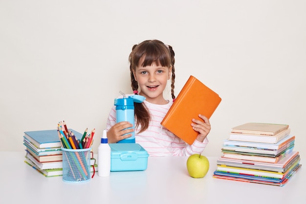 Inquadratura orizzontale di una piccola studentessa positiva con capelli scuri e trecce che tiene libro e bottiglia d'acqua blu seduta al tavolo circondata da libri contro il muro bianco