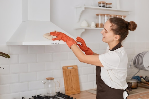 Photo horizontal shot of woman washing with a rag an electric cleaning hood at home kitchen wearing white t shirt brown apron and orange gloves housekeeper with bun hairstyle doing her work