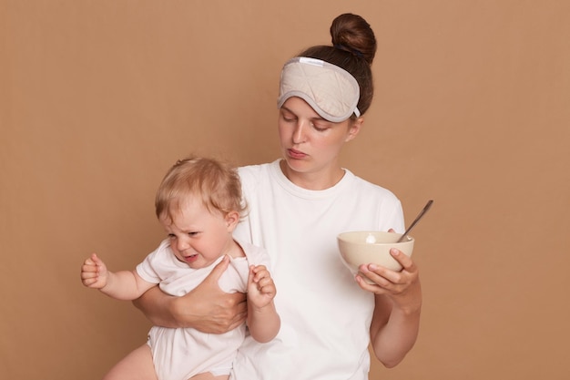 Horizontal shot of woman wearing white t shirt and sleeping mask mother holding crying baby daughter and plate with kid's food in hands posing isolated over brown background