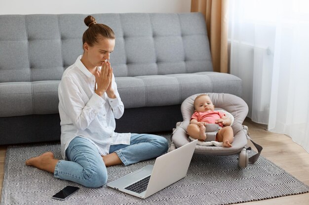 Horizontal shot of woman with bun hairstyle in white shirt and jeans working on notebook online and taking care of infant kid in rocking chair female posing with palms together in praying gesture